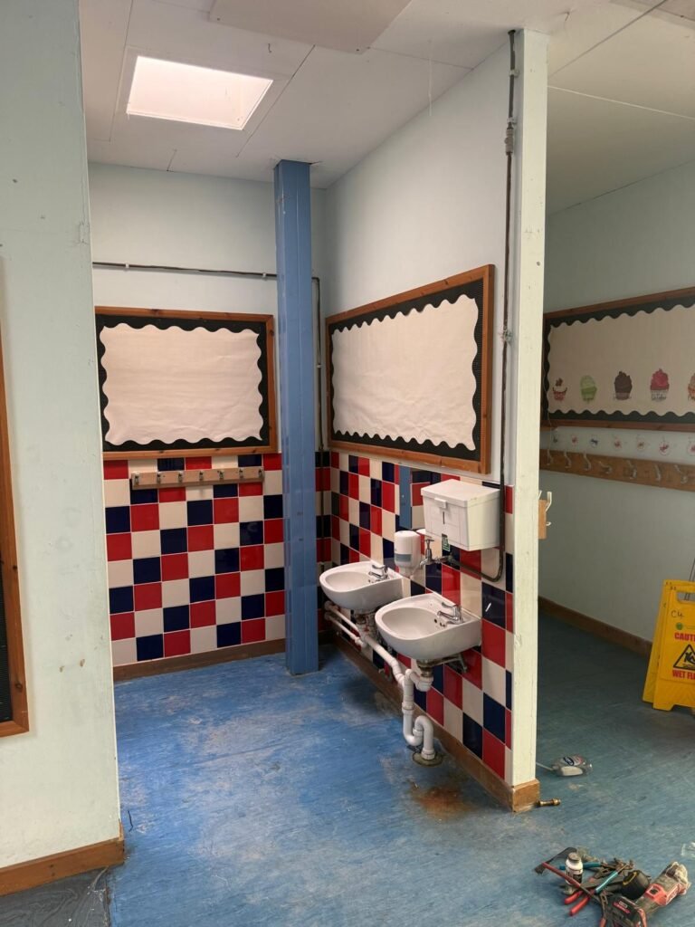 School washroom before refurbishment, with wall hung basins on red and blue chequerboard tiling, exposed waste pipes and a blue painted steel column