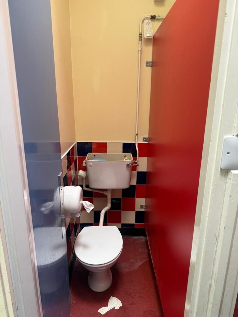 School toilet cubicle before refurbishment, with red and blue chequerboard tiling, an exposed low level cistern and surface run pipework