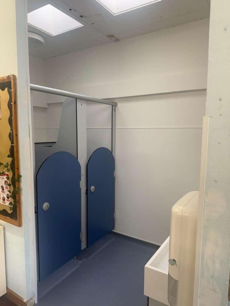 Run of finished school toilet cubicles with blue curved top doors and a grey support rail, beside a wash basin