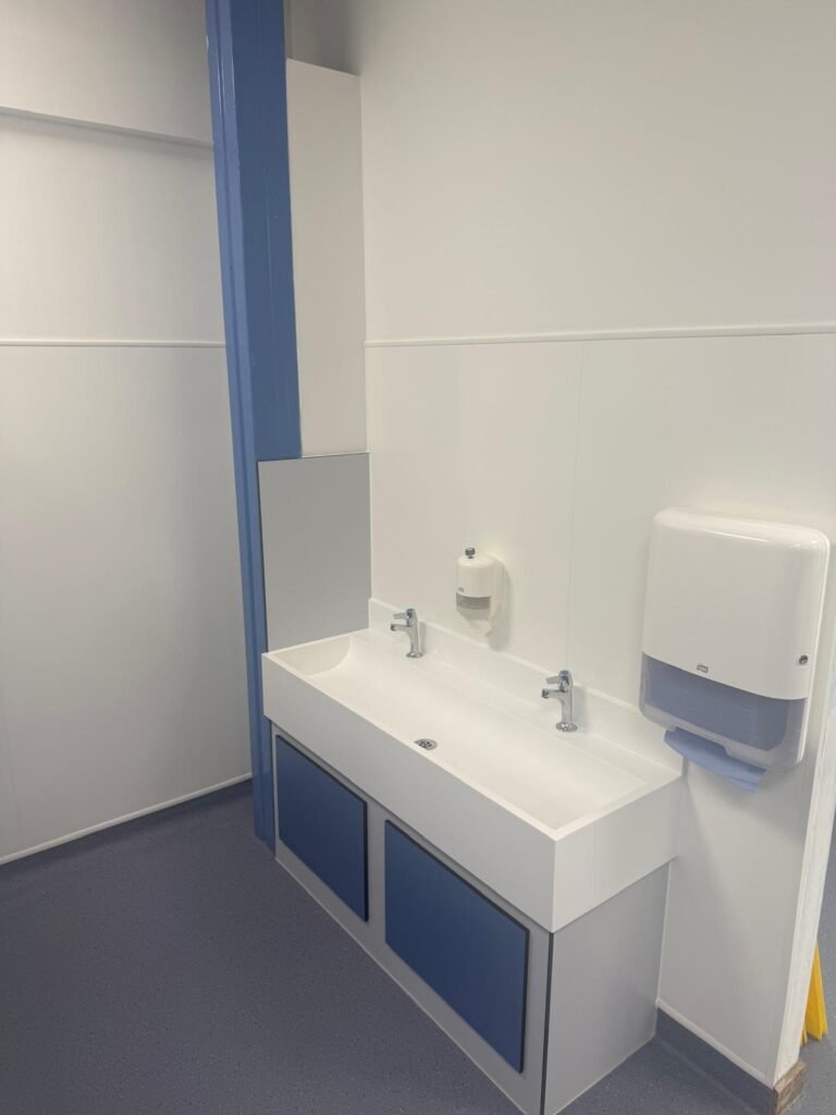 Finished school washroom with white Altro Whiterock wall cladding, blue safety vinyl flooring, a trough basin and a paper towel dispenser