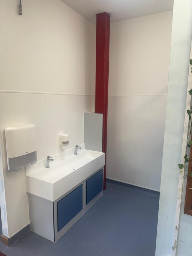 Finished school washroom with a solid surface trough basin on a panelled vanity unit, two taps, a mirror and full height wall panelling