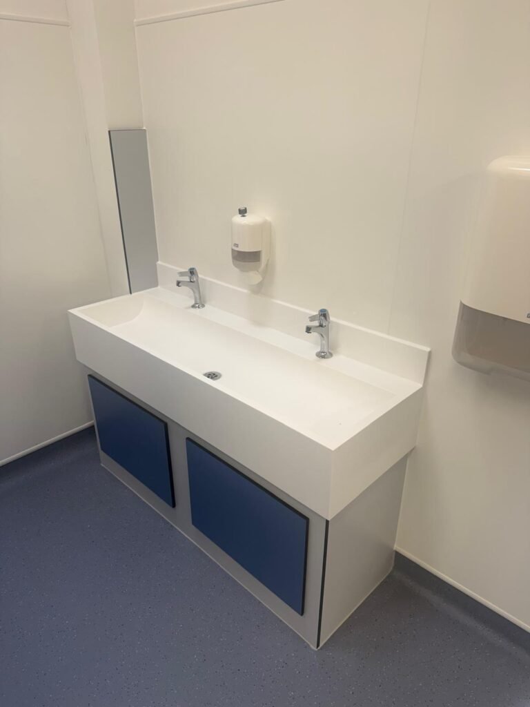 Finished school washroom vanity unit with a solid surface trough basin, two taps, a mirror and a soap dispenser against white wall cladding
