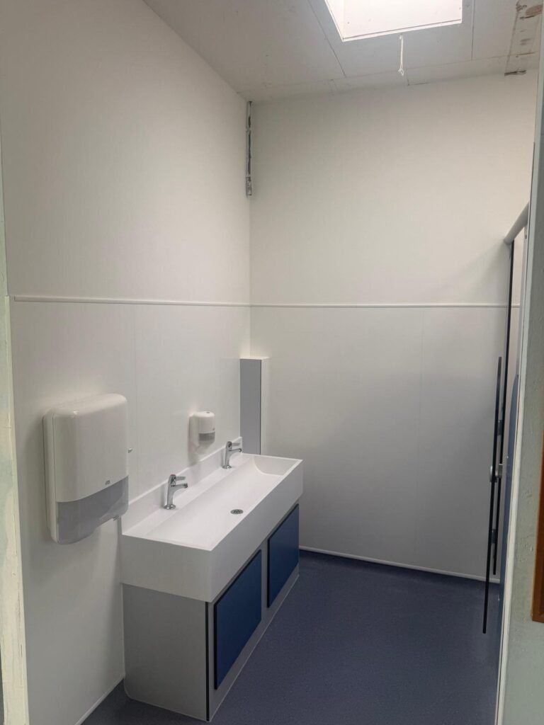 Finished school washroom with a solid surface wash trough and two taps on a blue panelled unit, white wall cladding and blue safety flooring
